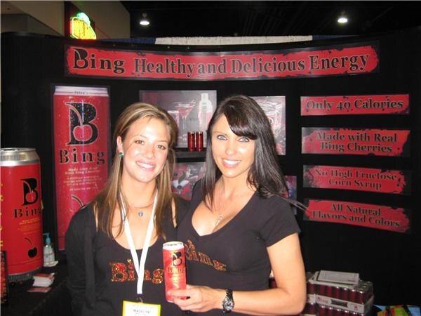 Two women in black Bing shirts smile in front of a red Bing booth display reading Healthy and Delicious Energy; the woman on the right holds a can of Bing.