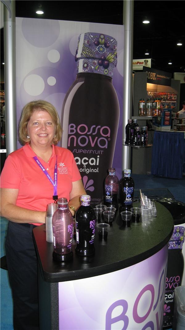 A smiling woman in a coral Bossa Nova polo stands at a curved counter with bottles of Bossa Nova açai juice and sample cups, in front of a large purple bottle banner.