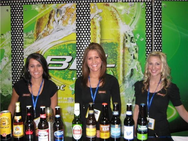 Three smiling women in black tops stand behind a counter lined with bottles including Leffe, Stella Artois, Widmer and Redhook ESB, in front of a Bud Light Lime backdrop.