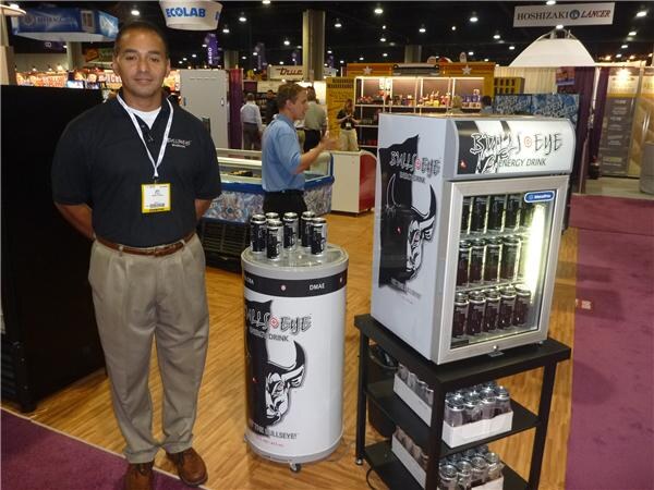 A man in a black polo and khakis stands beside a Bull's Eye Energy Drink mini cooler and a round display cooler topped with cans; another man talks in the background.