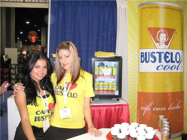 Two women in yellow Café Bustelo T-shirts stand by a small cooler of Bustelo Cool cans, with a tall Bustelo Cool can graphic on the right and sample cups on the table.