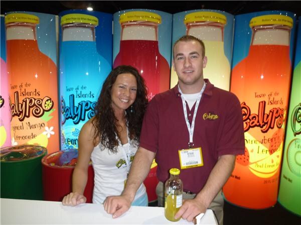 A smiling woman and a man in a maroon shirt stand at a counter in front of large lit Calypso lemonade bottle graphics; the man holds a bottle of Calypso.