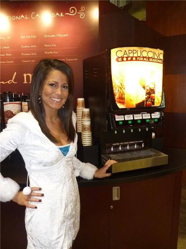 A smiling woman in a white fuzzy-trimmed jacket stands beside a lit Cappuccino For All Seasons dispenser with stacked paper cups and a flavor calendar on the wall.
