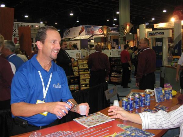 A smiling man in a blue Celsius polo holds a can and a cup at a booth table with Celsius cans and brochures; other attendees stand in the aisle behind him.