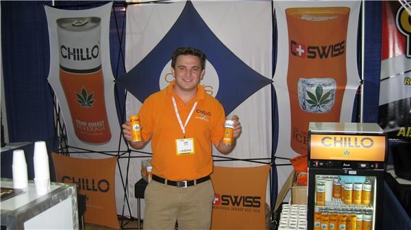 A smiling man in an orange polo and lanyard holds two cans in a booth with Chillo and Swiss hemp drink banners and a lit Chillo cooler filled with cans.
