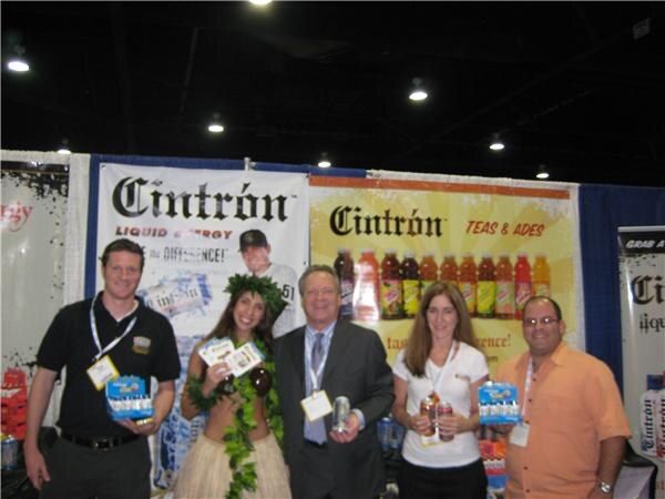 Five people pose in front of Cintrón banners; one woman wears a leaf costume, and some hold packages or cans. Bottles of Cintrón teas and ades line the backdrop.