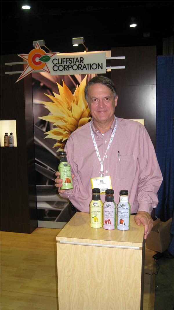 A man in a pink checked shirt holds a bottle of Chadwick Bay tea beside three more Chadwick Bay bottles on a wooden counter under a Cliffstar Corporation sign.