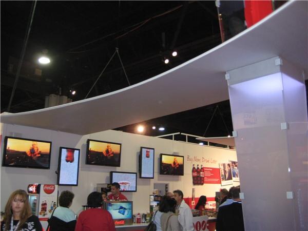 Wide view of a Coca-Cola booth with a curved white canopy overhead, video screens along the back wall and visitors standing at a counter in the foreground.
