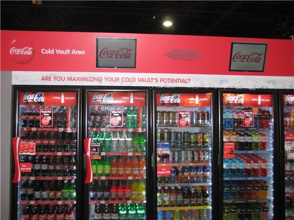 Row of glass-door Coca-Cola coolers stocked with bottled and canned drinks under a red banner reading Cold Vault Area and asking if you are maximizing your cold vault's potential.