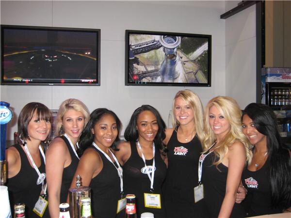 Seven smiling women in black tops and lanyards stand behind a bar with Coors Light and Miller Lite logos. Two TV screens hang on the wall behind them.