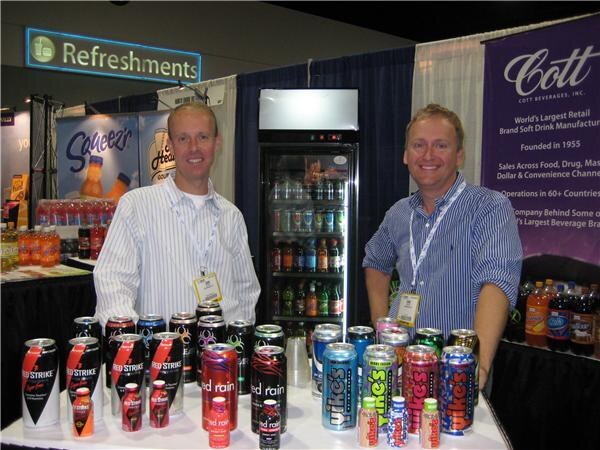 Two smiling men in striped shirts stand behind a table of Red Strike and Red Rain cans and other drinks, with a Cott banner and a Squeez sign behind them.