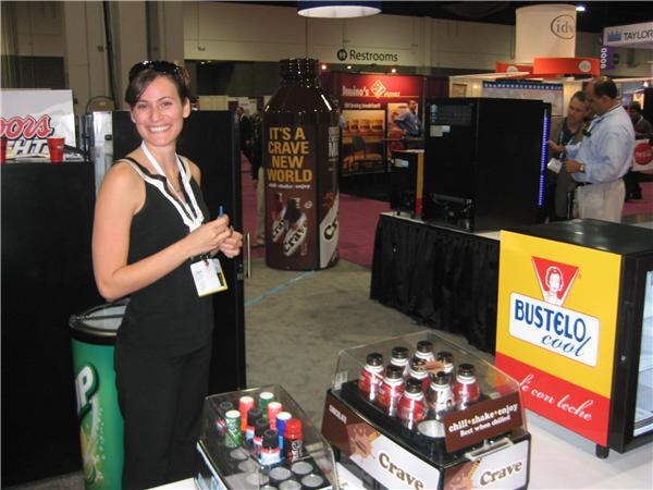 A smiling woman with a lanyard stands beside a counter with Crave bottles and an energy shot display. A Bustelo Cool cooler and a giant Crave bottle sign are behind her.