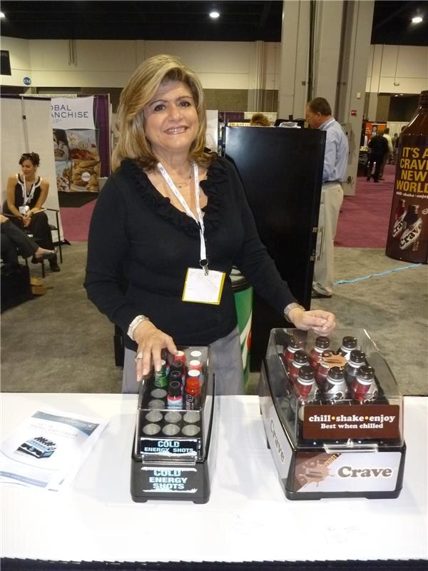 A smiling woman with a lanyard stands at a table with a Crave bottle display and a Cold Energy Shots display. A large Crave bottle-shaped sign is at the right.