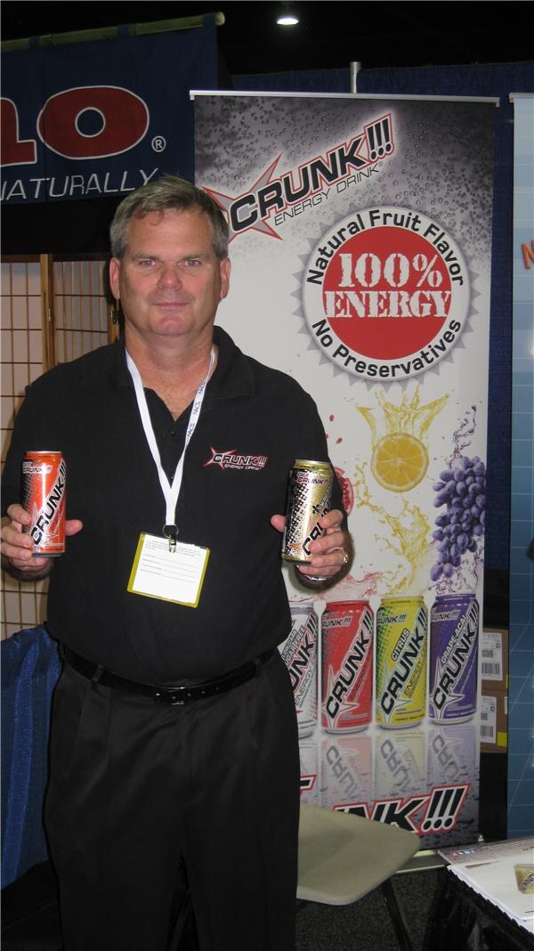 A man in a black polo and lanyard holds two Crunk!!! Energy Drink cans in front of a banner reading Natural Fruit Flavor, 100% Energy, No Preservatives.