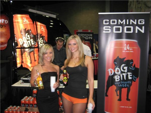 Two women hold beer bottles beside a Coming Soon banner showing a Dog Bite High Gravity Lager can. A man wearing headphones stands behind them near a black vehicle with Dog Bite graphics.