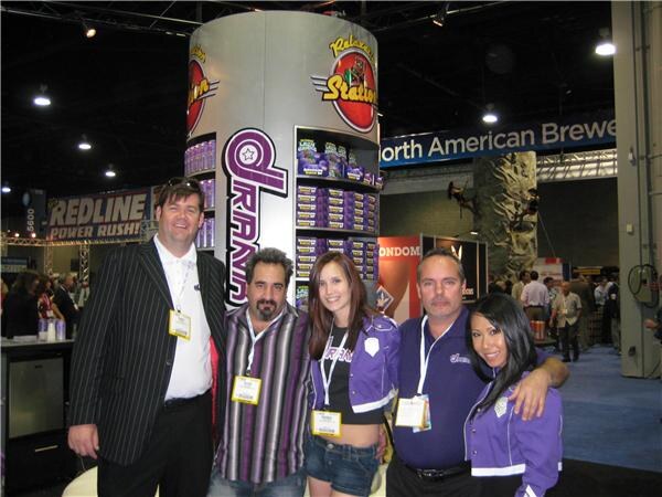 Five people in a group pose in front of a tall Drank display tower stocked with boxes; two women wear purple Drank jackets. A Redline Power Rush banner hangs in the background.