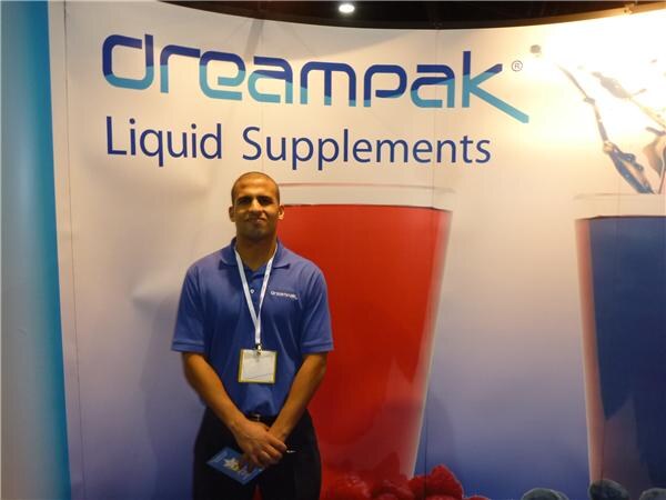 A man in a blue Dreampak polo with a lanyard and badge stands in front of a Dreampak Liquid Supplements backdrop showing glasses of red and blue drink and berries.