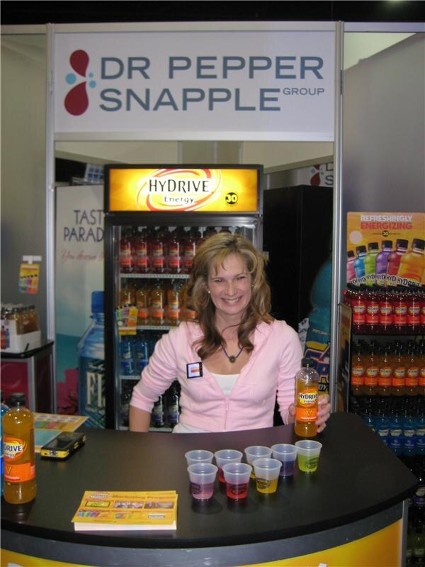 A smiling woman in a pink jacket holds a Hydrive Energy bottle behind a counter with sample cups, beneath a Dr Pepper Snapple Group sign and a lit Hydrive Energy cooler.