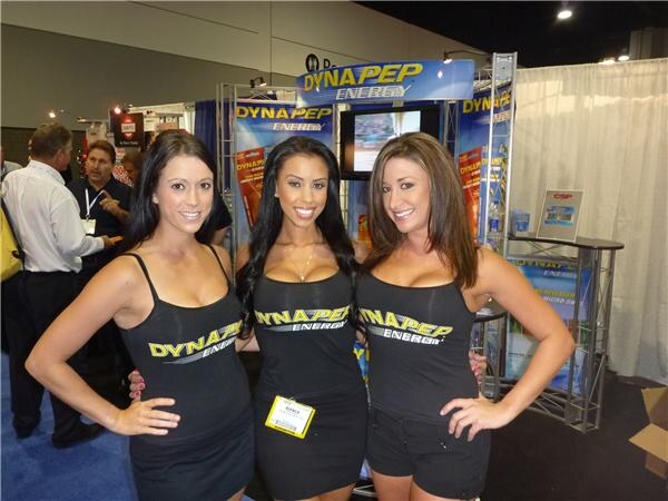 Three women in black Dynapep Energy outfits pose together in front of the Dynapep Energy booth, with its signs and a monitor behind them. Other attendees stand at left.