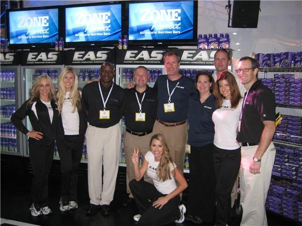 A group of nine men and women pose in front of EAS refrigerated displays stocked with product, with ZonePerfect screens above. One woman crouches in front making a peace sign.