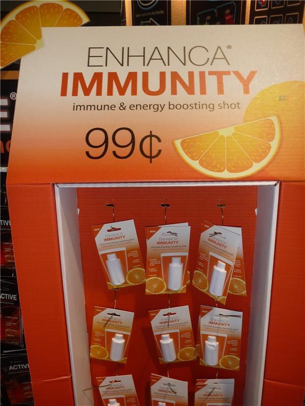 Orange cardboard display reading Enhanca Immunity, immune & energy boosting shot, 99¢, with orange slice graphics and rows of hanging packs, each holding a small white bottle.