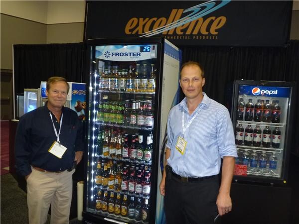 Two men with lanyards stand beside a lit Froster glass-door cooler filled with bottles, under an Excellence Commercial Products sign. A Pepsi cooler stands at right.
