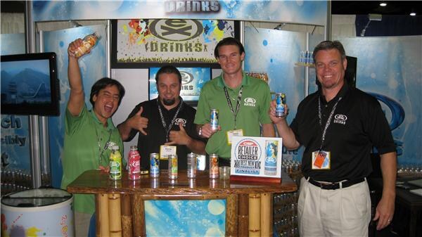 Four men in Ex Drinks shirts stand behind a bamboo-style counter holding cans and bottles; one raises a bottle. A Retailer's Choice finalist sign sits on the counter.