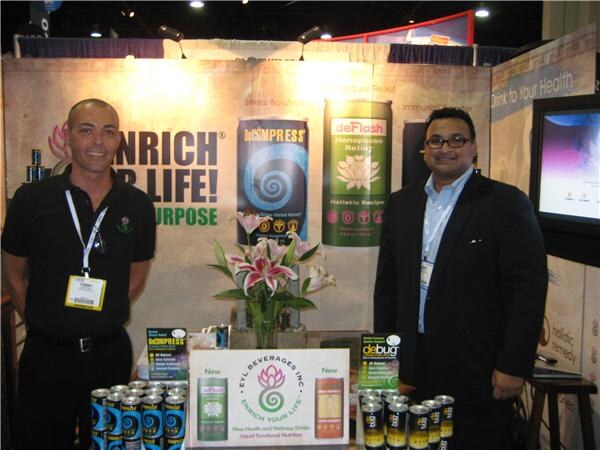Two men stand at the EYL Beverages booth with lilies on a table and stacked cans of DeCompress, deFlash and deBug. Banners behind them show the products.