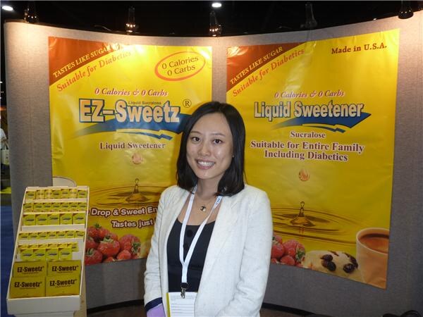 A smiling woman with a lanyard stands in front of yellow EZ-Sweetz liquid sweetener banners, with a display of EZ-Sweetz boxes at left.