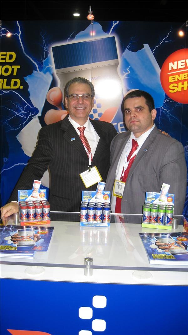 Two men in suits and red ties stand behind a display counter of X-Force energy shot cans. The man on the right wears a badge reading Fabricio. A lightning-themed backdrop is behind them.