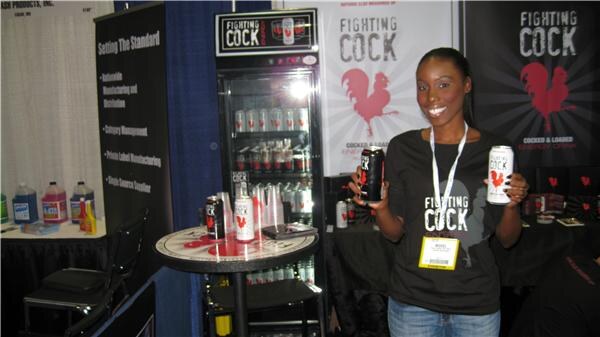 A smiling woman in a black Fighting Cock T-shirt and badge holds two cans of Fighting Cock energy drink beside a lit cooler and rooster-logo banners.