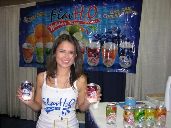 A smiling woman in a white Flav H2O tank top holds two canned drinks in front of a Flav H2O banner with fruit images; more cans sit on a table at right.