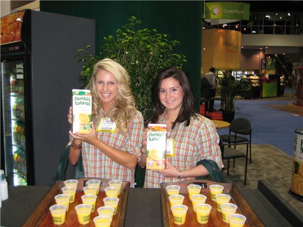 Two smiling women in plaid shirts each hold a carton of Florida's Natural orange juice behind trays of small cups filled with juice.