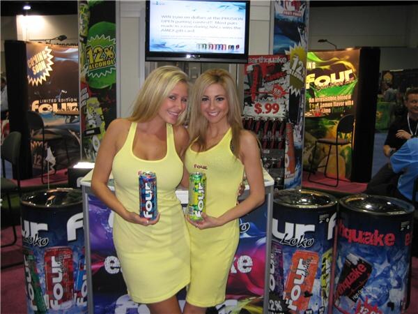 Two women in yellow dresses hold cans of Four Loko in front of a counter and barrel-style coolers with Four Loko and Earthquake graphics, with a Phusion Projects display behind.