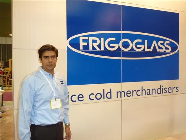 A man in a light blue shirt and badge stands beside a large blue Frigoglass sign reading ice cold merchandisers.
