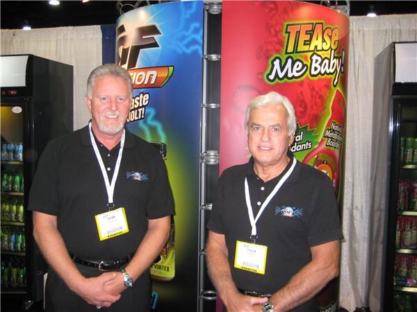Two men in black polo shirts and badges stand in front of Fusion and TEAse Me Baby! display columns, with beverage coolers on either side.