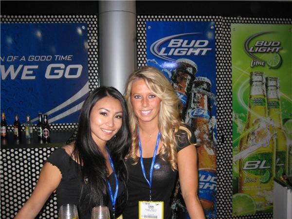 Two smiling women with lanyards pose in front of Bud Light and Bud Light Lime posters. The woman on the right wears a badge reading Gabi.