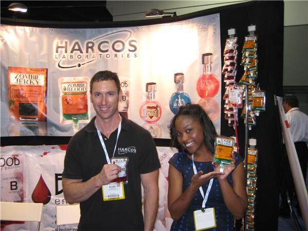 A man in a black Harcos shirt and a woman in a blue dress, both with badges, hold Zombie Blood and Zombie Jerky packages in front of the Harcos Laboratories banner.