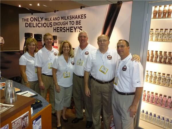 Five people in white polo shirts and badges stand together in front of a Hershey's milk and milkshakes banner, beside a lit cooler of bottled Hershey's milk drinks.