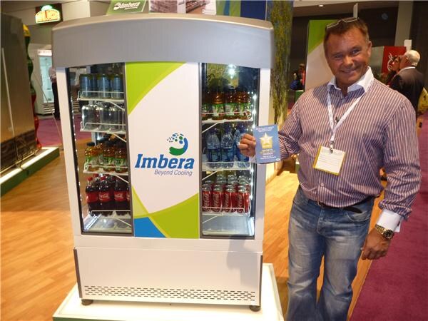 A smiling man with a lanyard badge holds a small card beside an Imbera display cooler stocked with Coca-Cola and other bottled and canned drinks.
