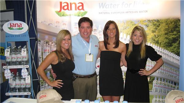 A man in a light blue shirt stands between three women in black dresses at a Jana booth with Water for Life signs and shelves of bottled Jana water behind them.