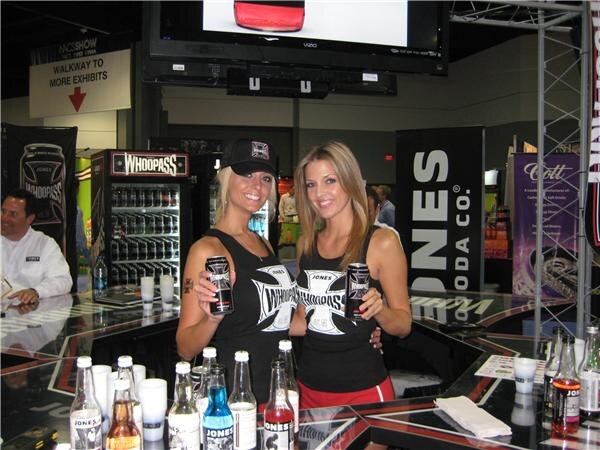 Two women in black Jones Soda tops hold cans of Whoopass at a counter lined with Jones Soda bottles, with a Whoopass cooler and Jones Soda Co. banner behind them.