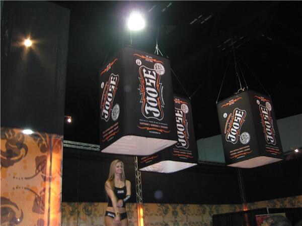 Three black box-shaped Joose banners hang from the ceiling over a trade show floor, with a woman in a black top standing below them.