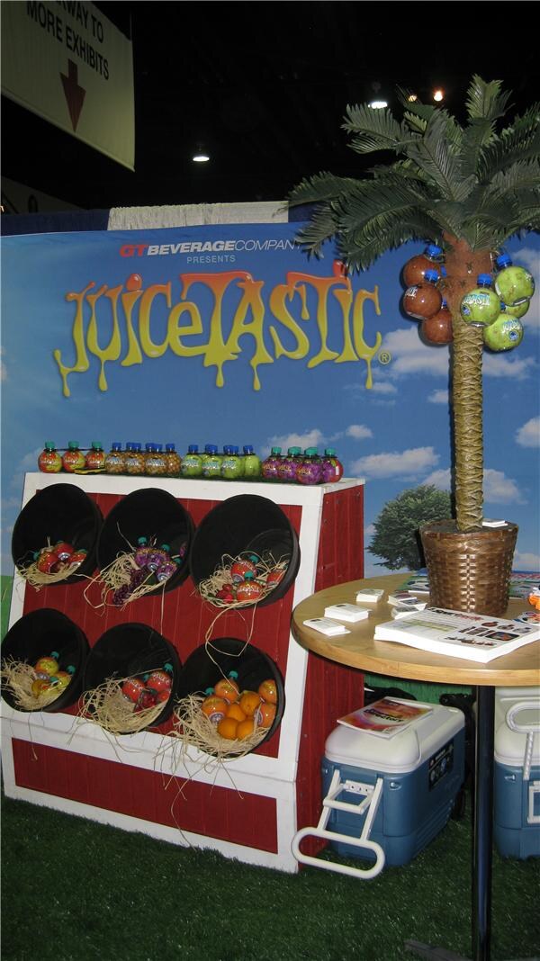 A Juicetastic display from GT Beverage Company with a barn-style shelf holding bottles in black buckets, a palm tree hung with bottles, and a table with brochures and coolers.