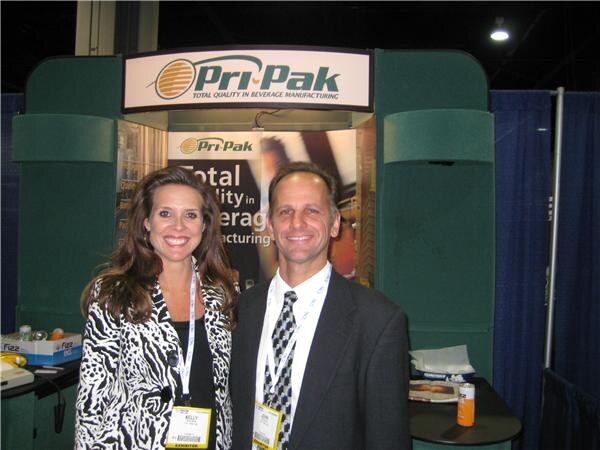 Kelly, on the left in a zebra-print jacket and exhibitor badge, stands beside a man in a dark suit and tie at the Pri-Pak booth.