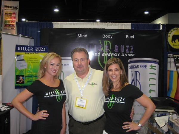 A man in a pale yellow polo stands between two women in black Killer Buzz shirts in front of a Killer Buzz sustained energy drink banner.