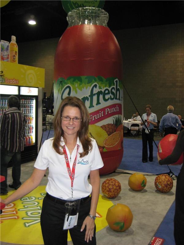 A woman in a white La Croix polo and red lanyard stands in front of a giant inflatable Everfresh Fruit Punch bottle, with inflatable fruit on the floor and a cooler to the left.