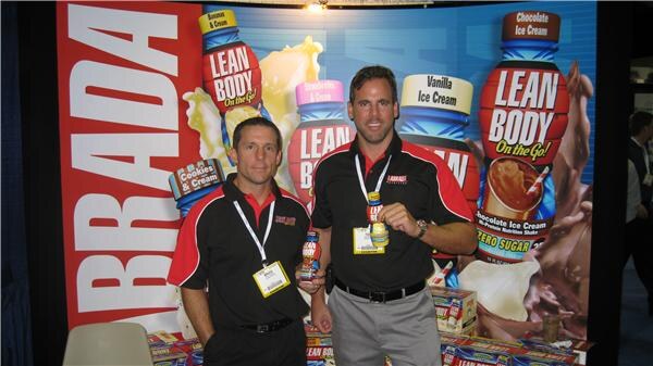 Two men in black and red polo shirts with lanyards stand in front of a Lean Body display of bottles; one holds a small bottle. Boxes of product sit in front of them.