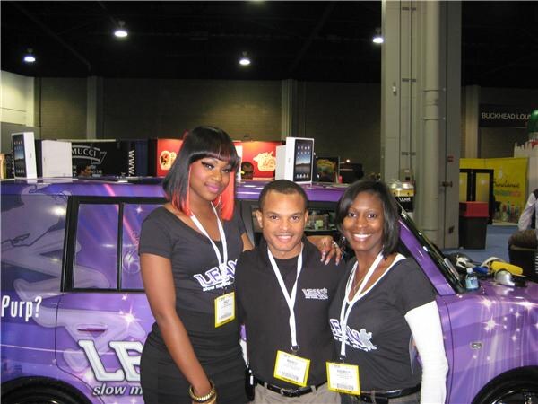 Two women and a man in black Lean slow motion shirts and badges pose in front of a purple vehicle with Lean graphics, inside a convention hall.