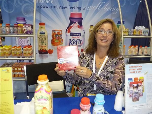 A woman with glasses holds a red Lifeway Kefir bar box at a table with Lifeway Kefir and LaFruta bottles, in front of a display of Lifeway kefir bottles.